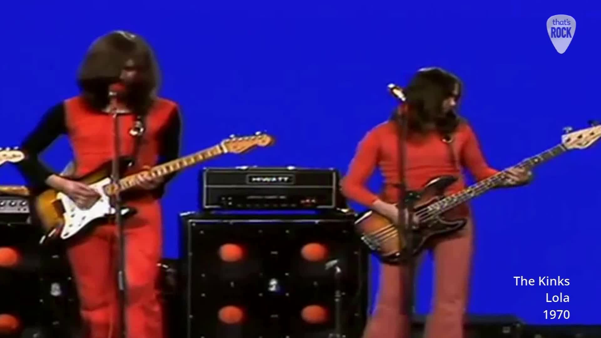 A guitarist in a vibrant orange jumpsuit plays a sunburst Stratocaster. Beside him, a bassist in a matching outfit moves his fingers across the fretboard of a dark bass guitar. A guitarist in a vibrant orange jumpsuit plays a sunburst Stratocaster. Beside him, a bassist in a matching outfit moves his fingers across the fretboard of a dark bass guitar.