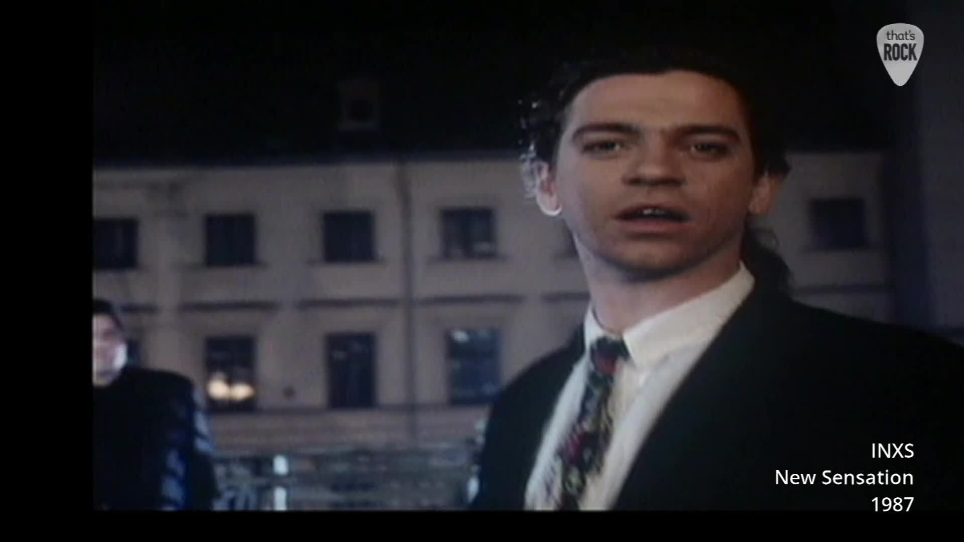 A man in a suit and tie turns his head, his expression caught mid-sentence. Behind him, another figure walks past the dark windows of a building. A man in a suit and tie turns his head, his expression caught mid-sentence. Behind him, another figure walks past the dark windows of a building.