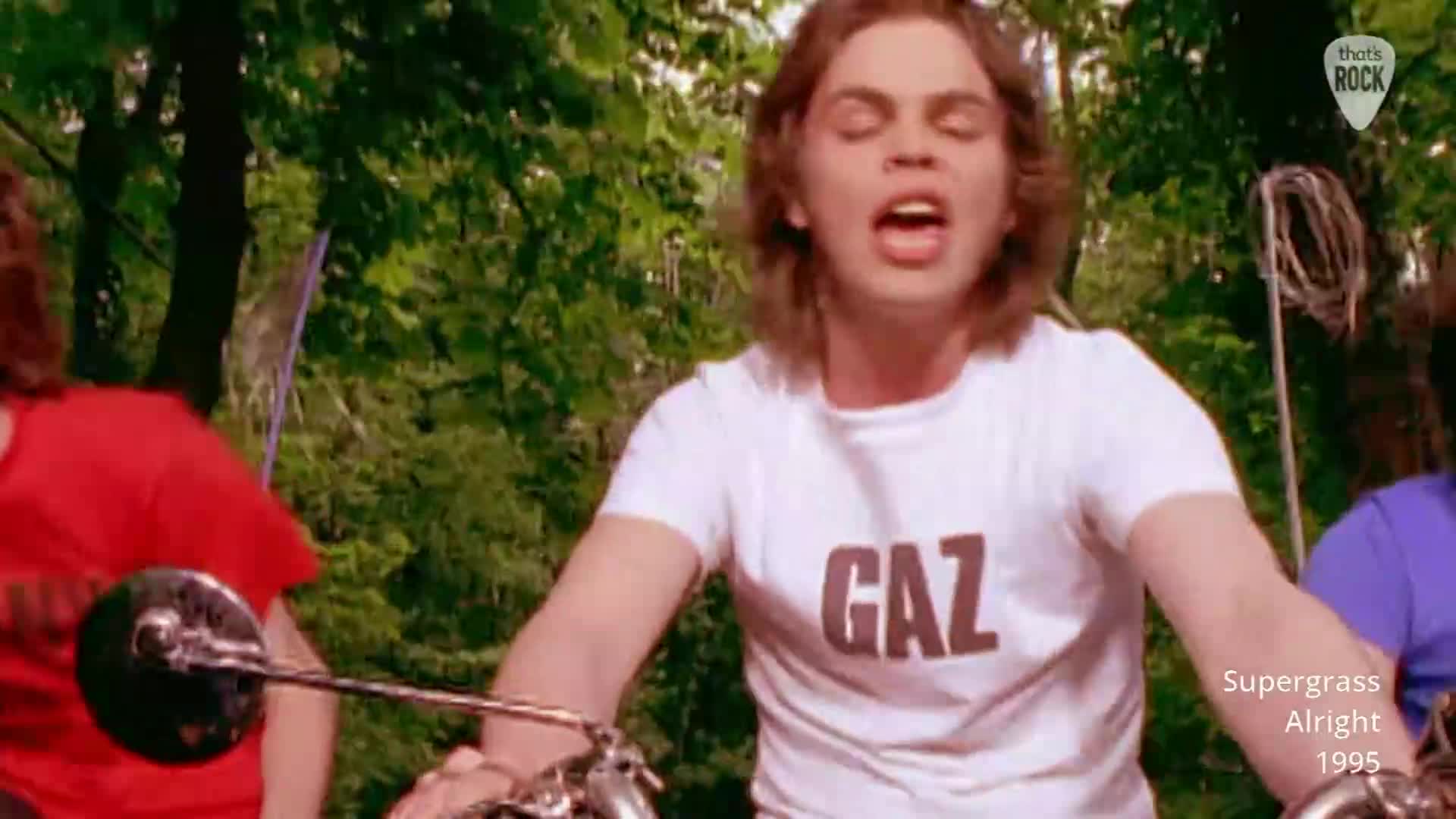 A young man with long hair, wearing a white t-shirt with "GAZ" printed on it, is riding a motorbike. He's singing with his mouth wide open, and a person in a red shirt is visible to his left.
