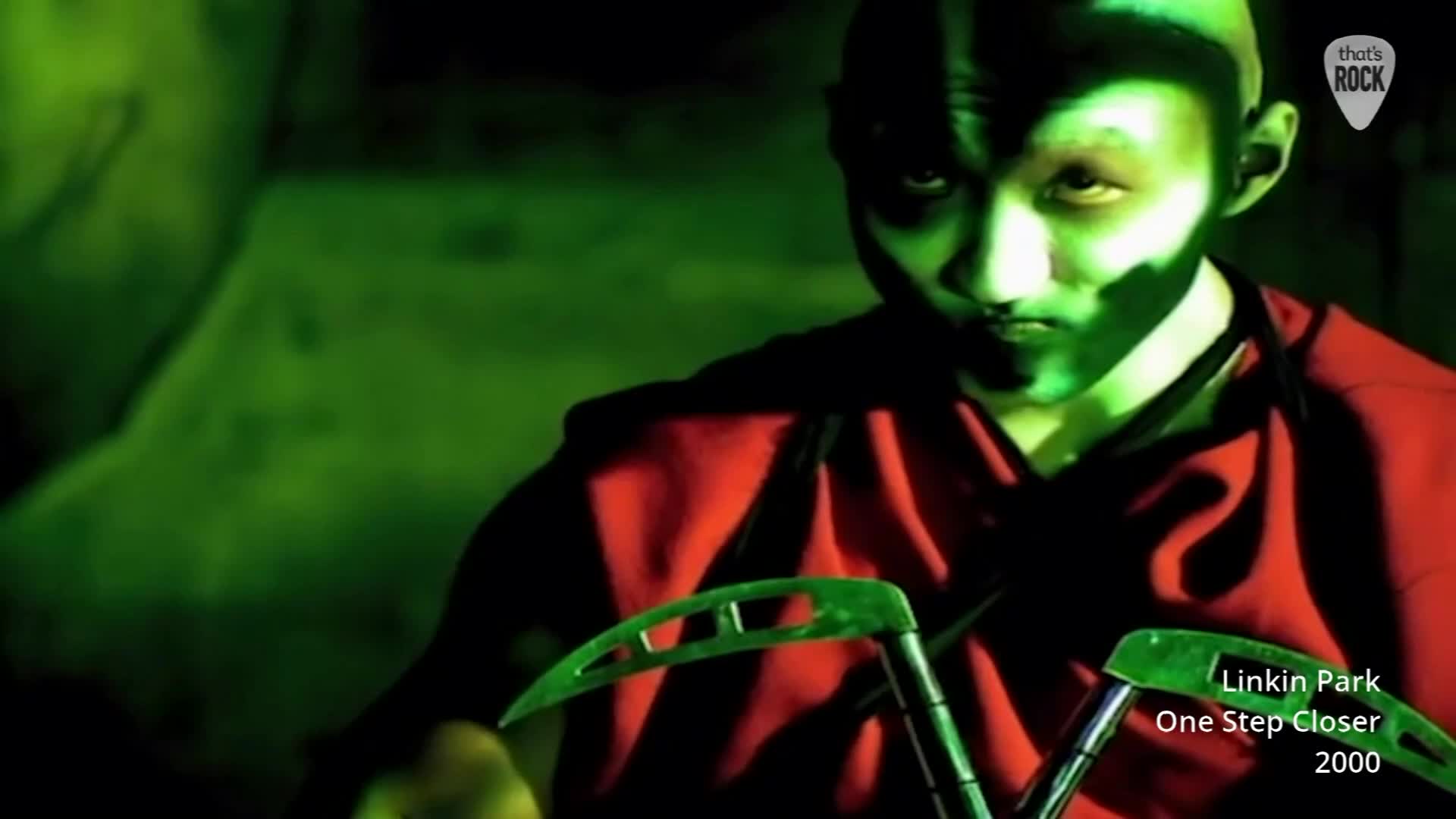 A figure in a red robe grips two scythe-like weapons, their face illuminated by a stark green light. The intense glow casts sharp shadows across their determined expression.
