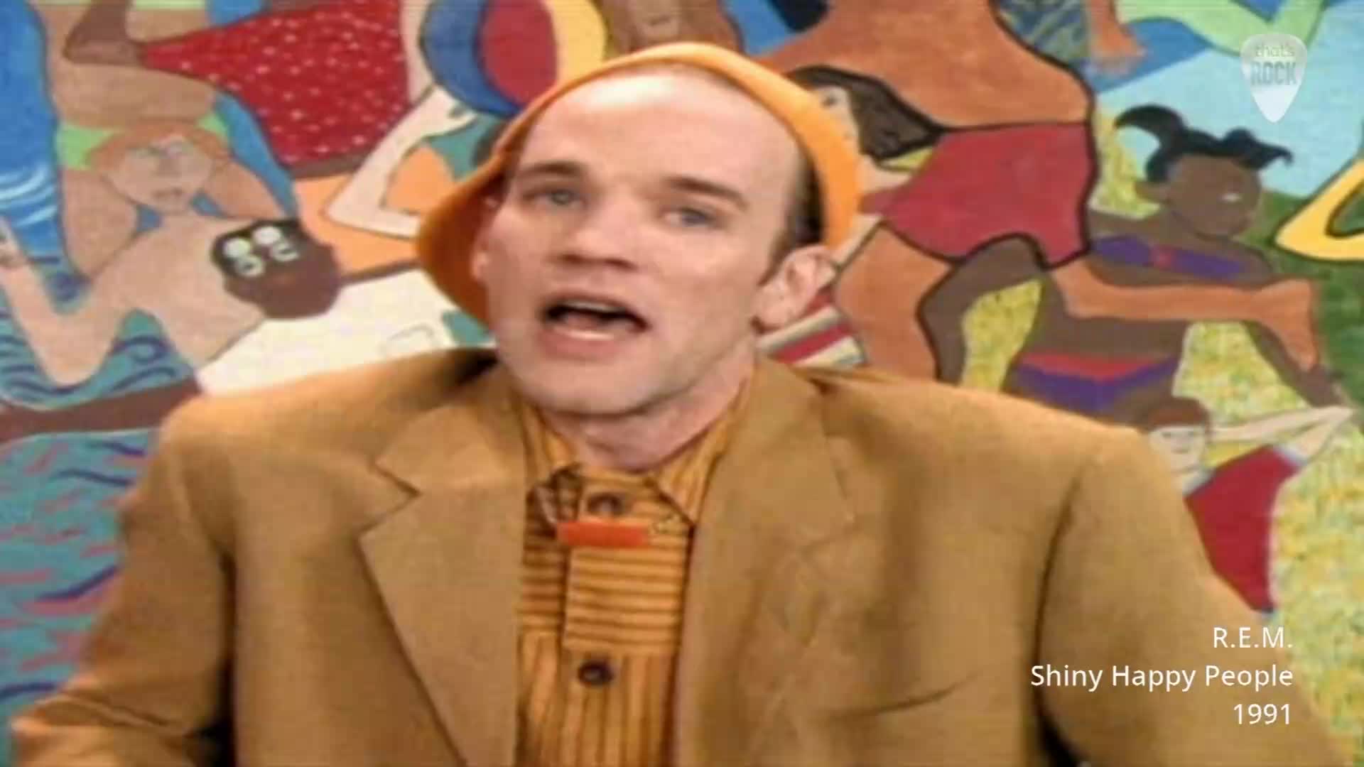 The man in the orange hat sings into the microphone, his mouth open. Behind him, a colourful mural depicts people in swimsuits running and playing.