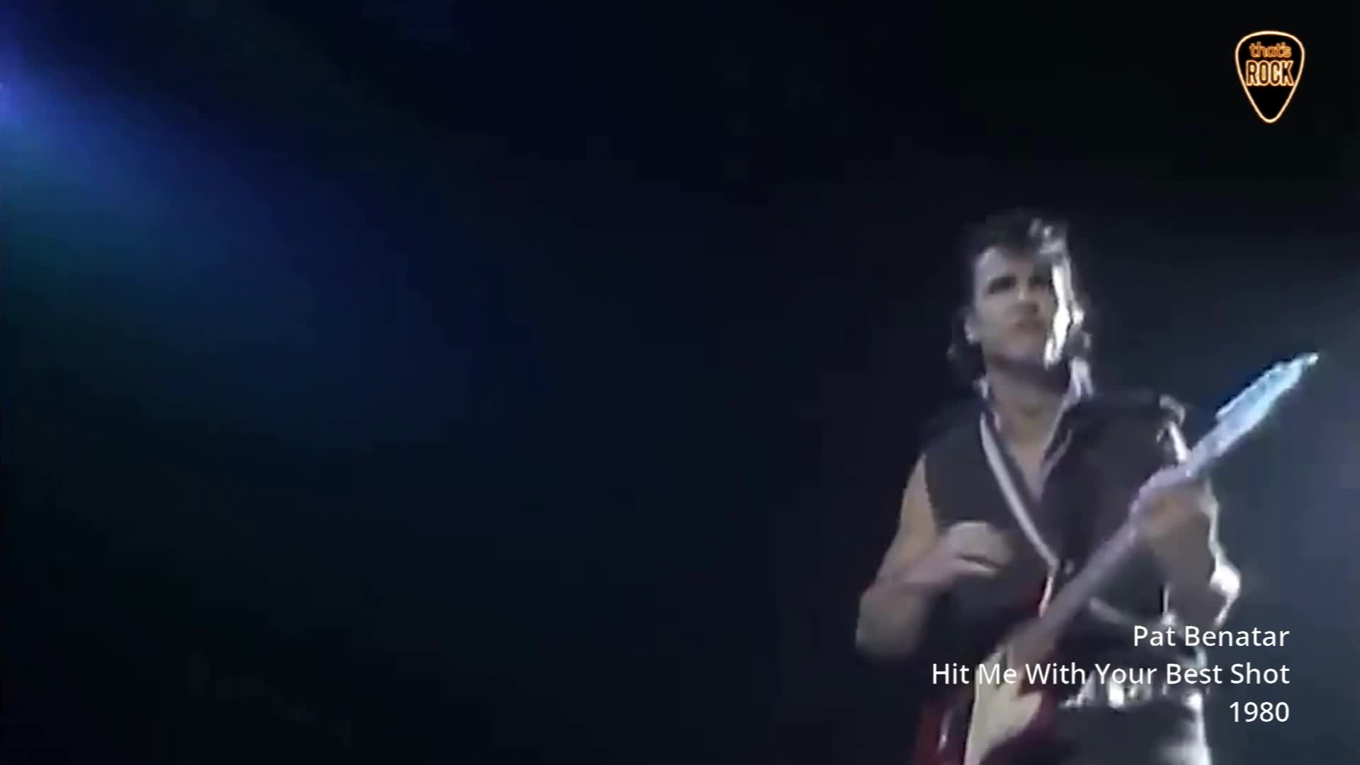 A guitarist in a dark vest plays a white electric guitar, his arm raised. A blue spotlight shines from the left, illuminating the stage.