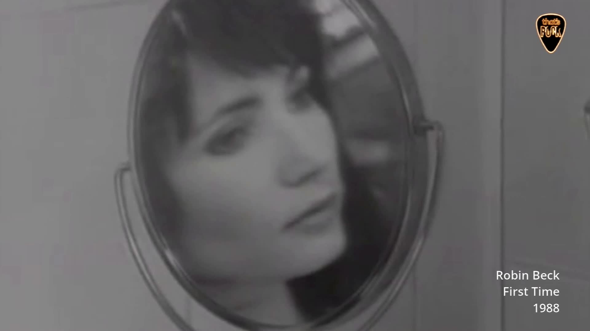 A woman's face is reflected in a round mirror, her gaze directed slightly away. The grainy black and white footage, likely from a music video from the late 80s, captures her thoughtful expression.