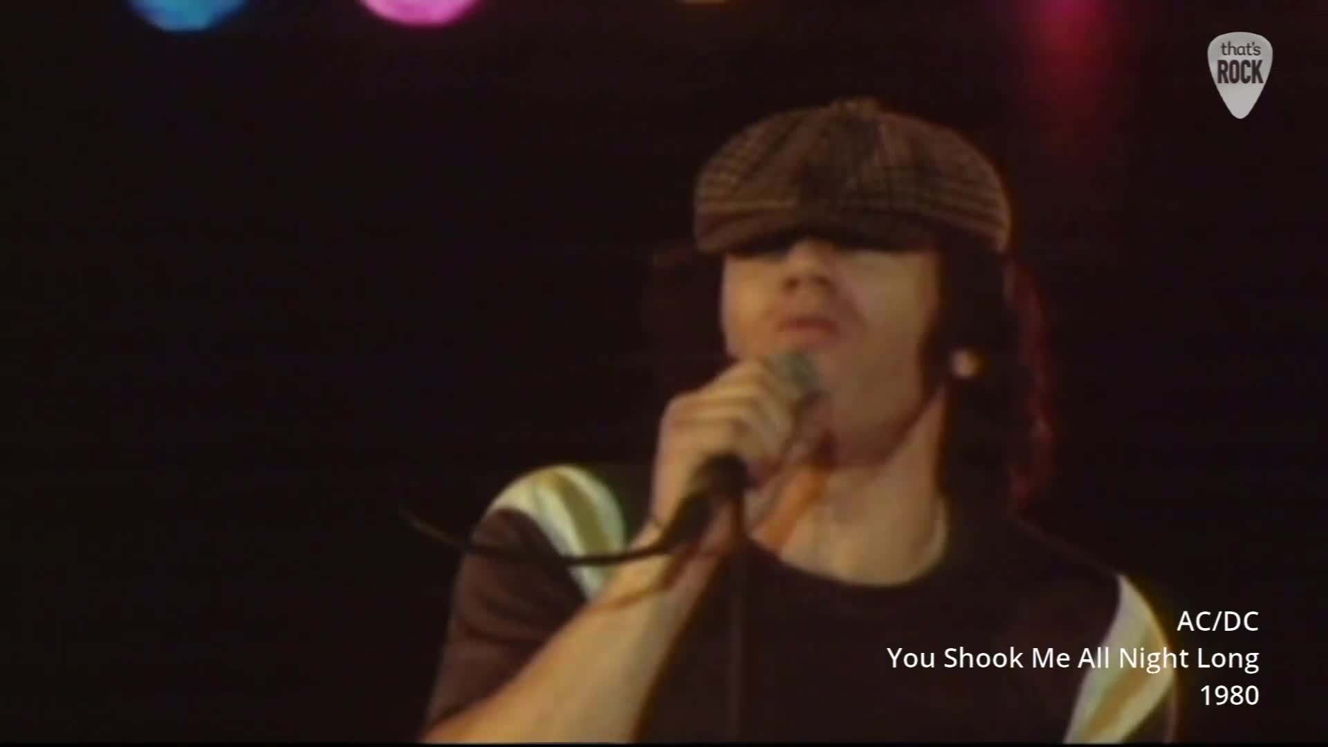 Bon Scott belts into the microphone, his flat cap pulled low. The lights above cast a dim glow on the stage.