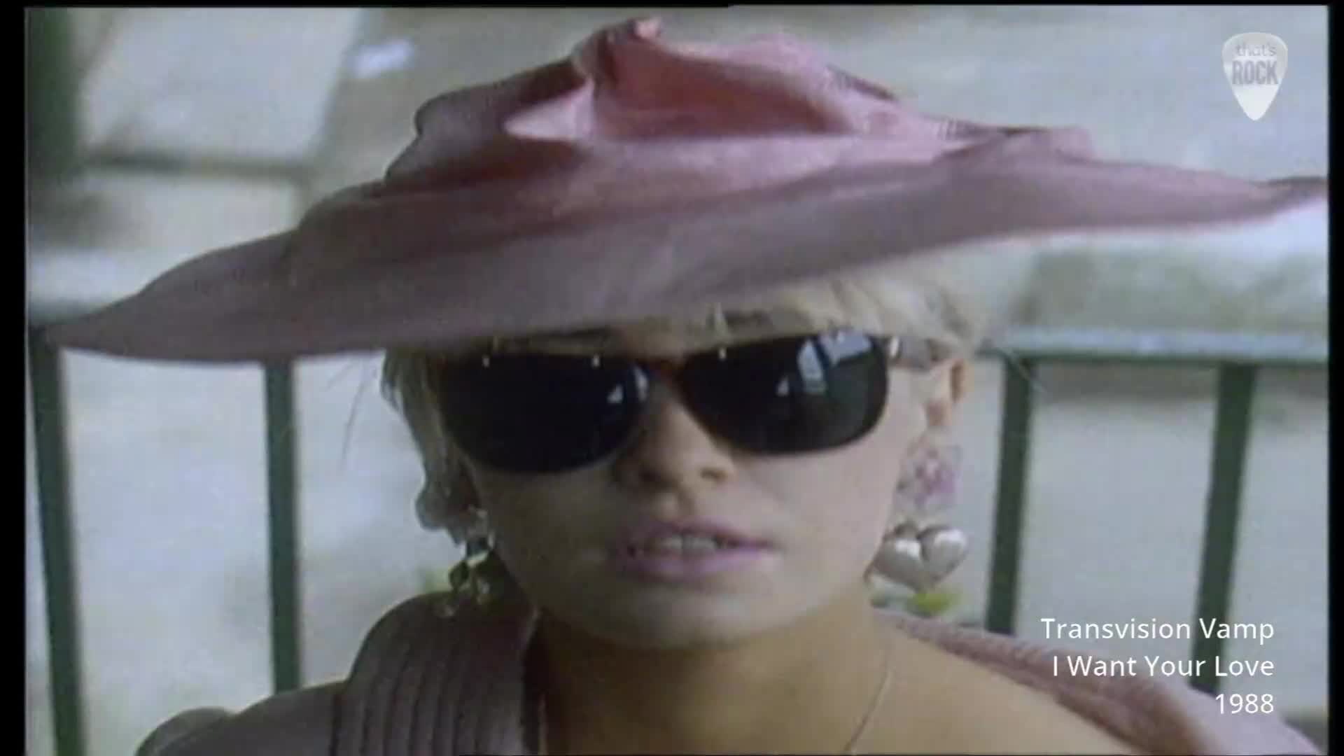 A woman with blonde hair, wearing large sunglasses and a wide-brimmed pink hat, looks directly ahead. Her large, heart-shaped earrings swing slightly as she moves.