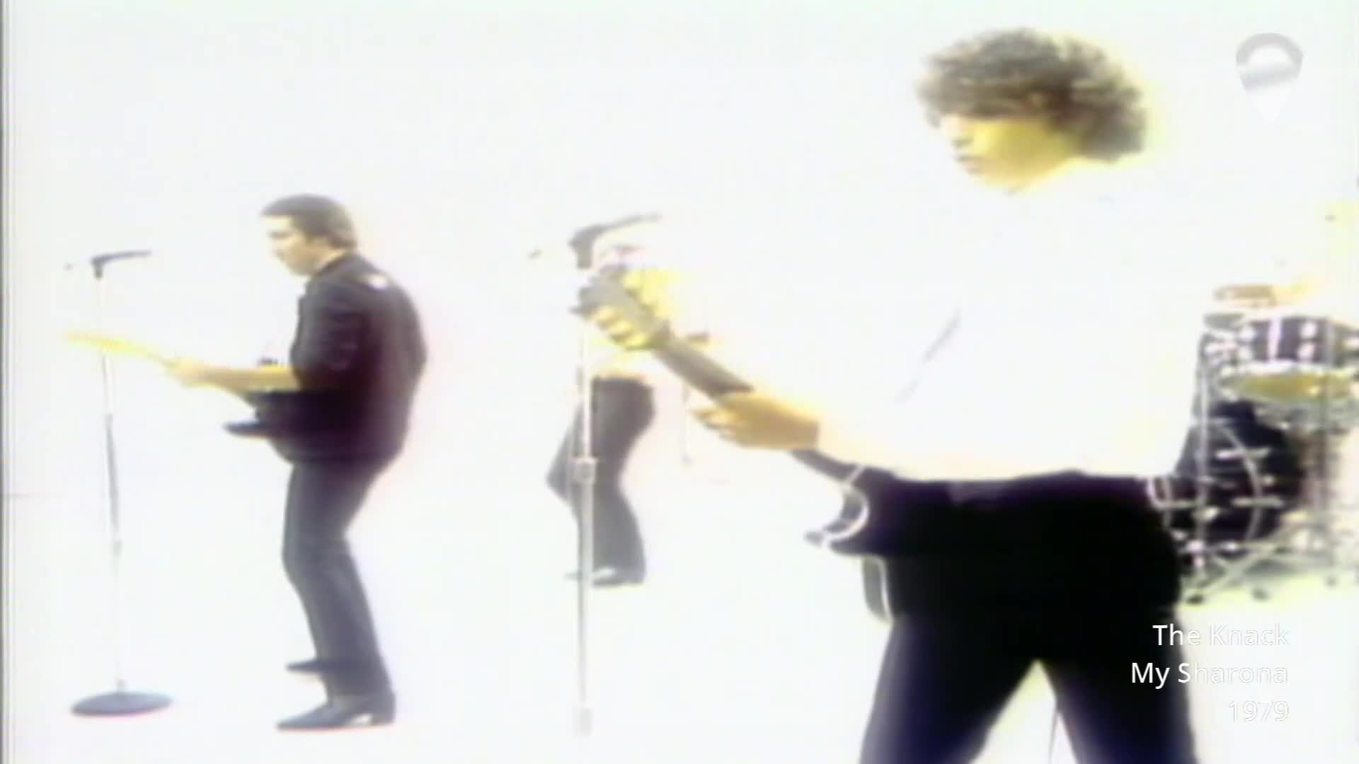 A guitarist in a black shirt plays his instrument, his body angled towards the audience. Another guitarist, dressed in white, stands to his right, his curly hair framing his face as he strums.