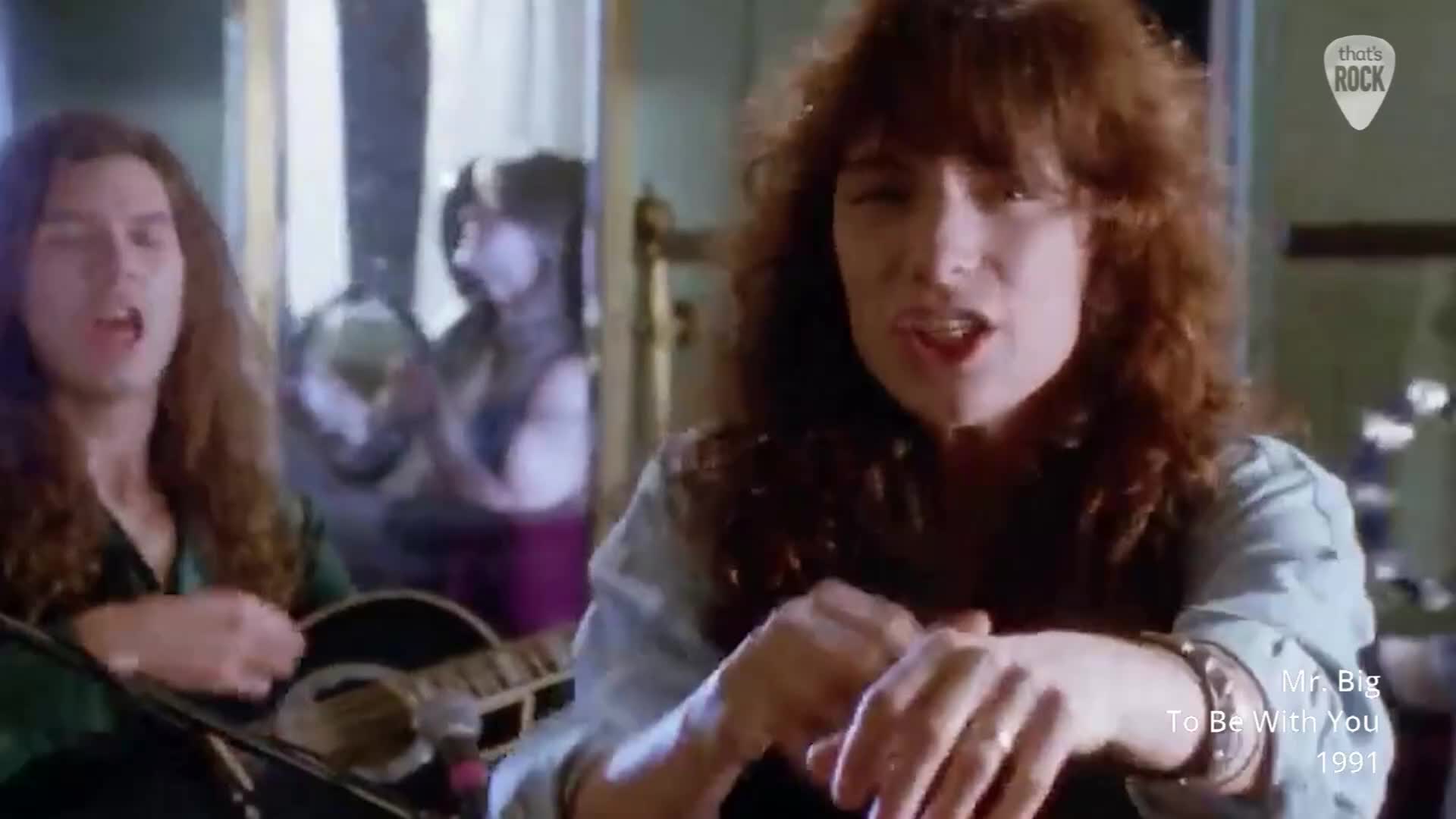 A man with long, brown hair sings into a microphone while playing an acoustic guitar. Another man, closer to the camera, is also singing, his hand resting on his arm.