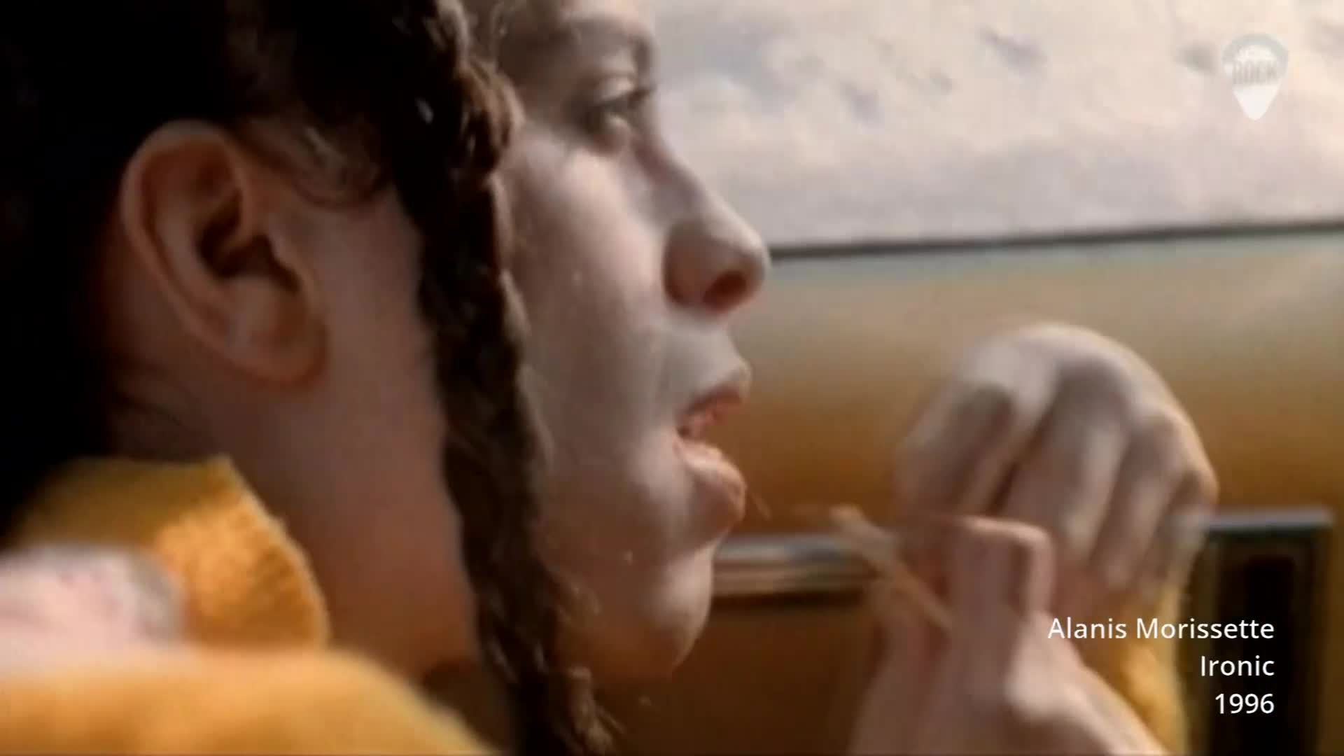 The girl is eating chips in the passenger seat of a car. Alanis Morissette's "Ironic" plays, a hit from 1996.