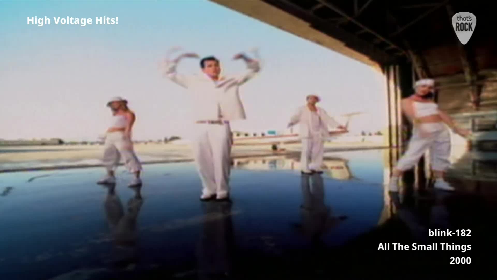 Four people in white outfits are dancing in front of an open hangar. A plane sits behind them on the tarmac.