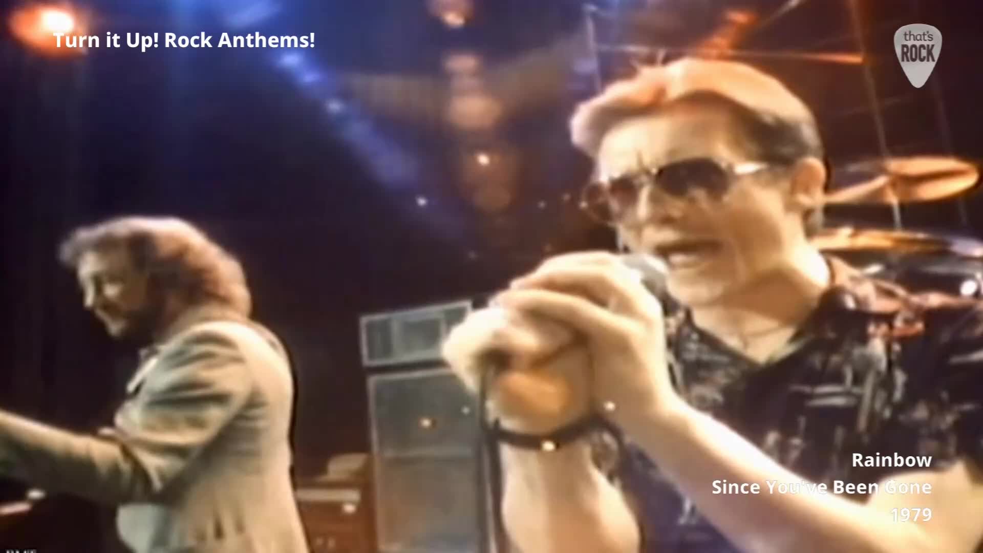 The singer, sunglasses on, grips the microphone, his mouth open as he belts out the lyrics. Beside him, a guitarist in a light jacket sways, his instrument held low.