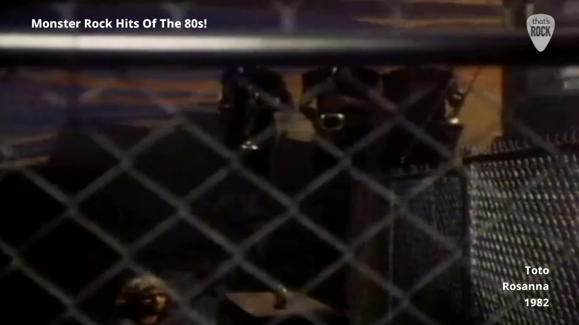 A band is on stage, bathed in dim light, with a drummer visible behind a kit. The performance is for a crowd, though they are obscured by a chain-link fence.