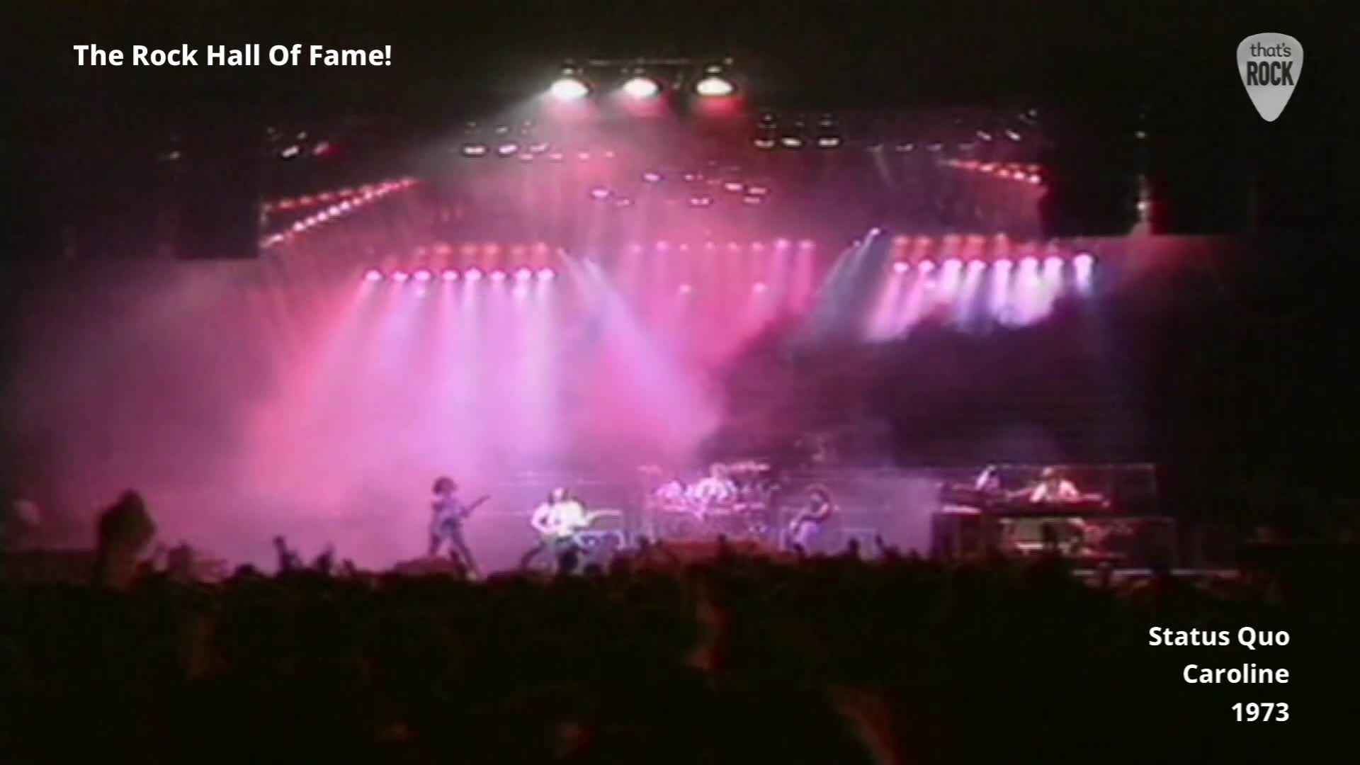 The band Status Quo are on stage, bathed in pink light. A drummer is visible behind his kit, and the guitarist on the left is mid-stride.