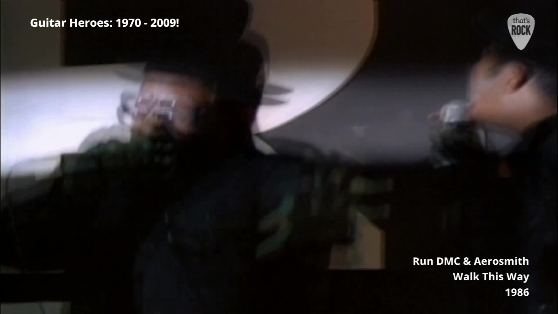 A man in glasses is singing into a microphone, his head tilted back. Another man, holding a microphone close to his lips, leans in from the right.