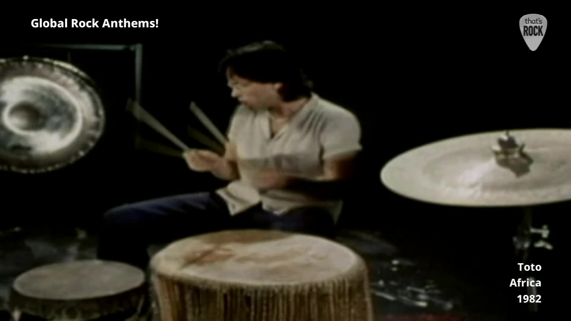 A drummer in a light shirt powerfully strikes his kit, sticks a blur against the dark backdrop. Large drums and a cymbal surround him as he plays.