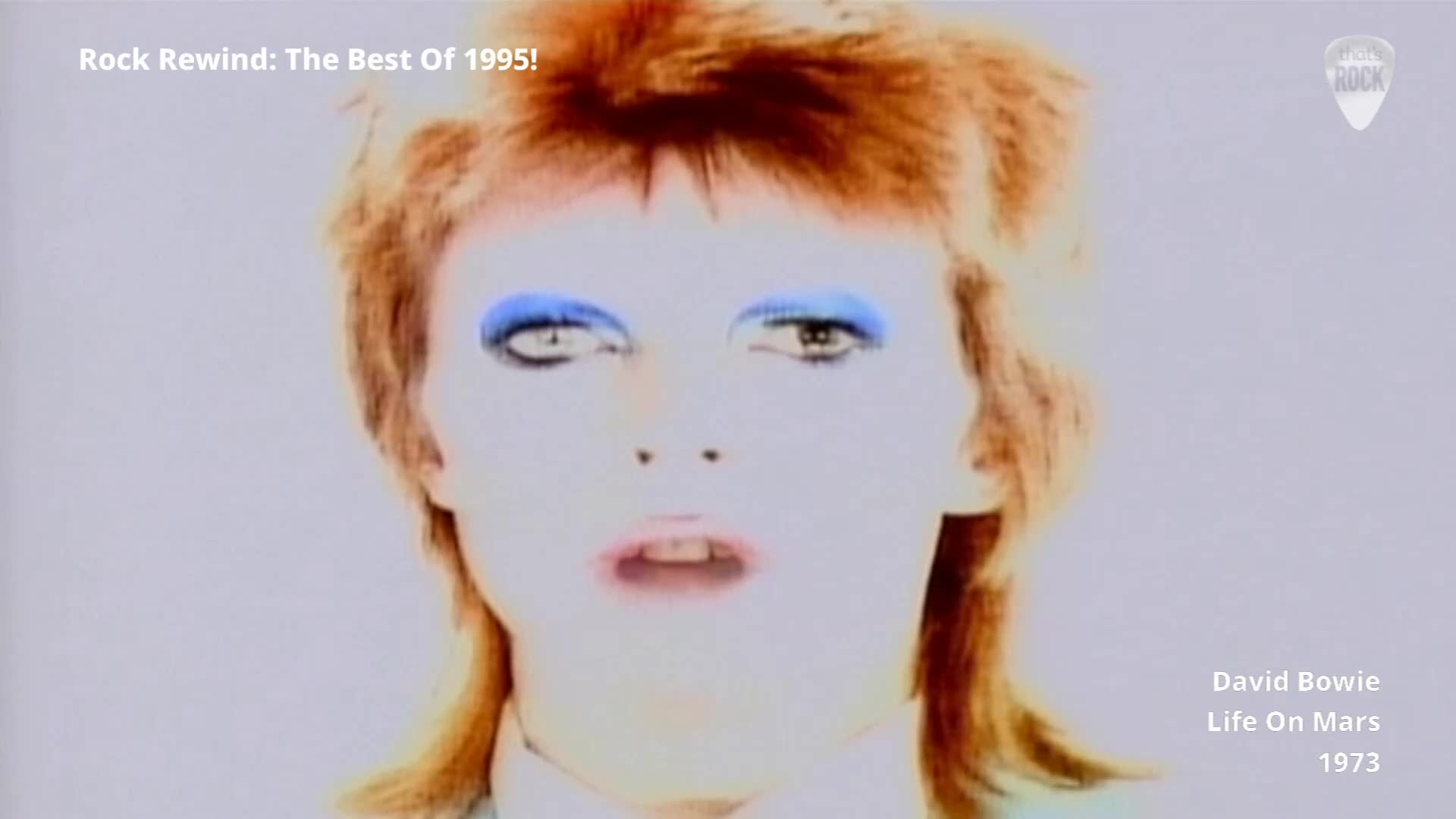 David Bowie's face fills the screen, his bright blue eyeshadow stark against pale skin. His mouth is slightly open, mid-sentence, as the camera focuses on his iconic look.