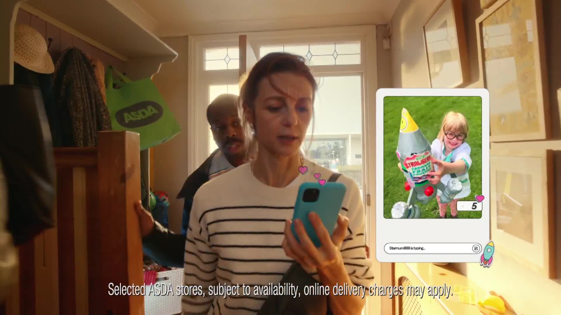 A woman in a striped top stares intently at her phone, a social media post of a child with a rocket balloon on the screen. Behind her, a man peers over her shoulder, and an Asda bag hangs on the coat rack.
