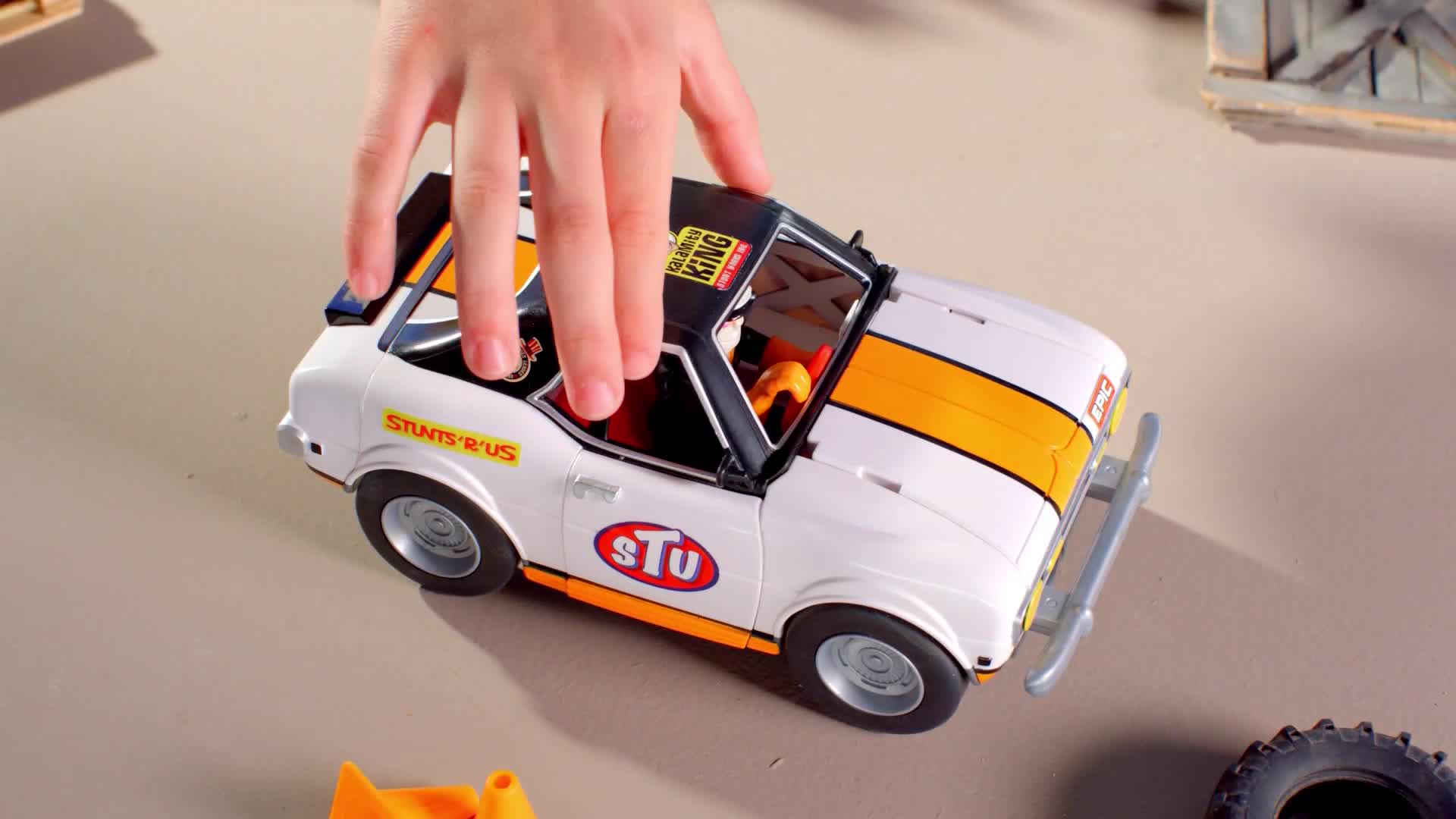 A child's hand hovers above a white toy car with orange racing stripes. The car, branded "Stu," sits on a light-colored surface, ready for play.
