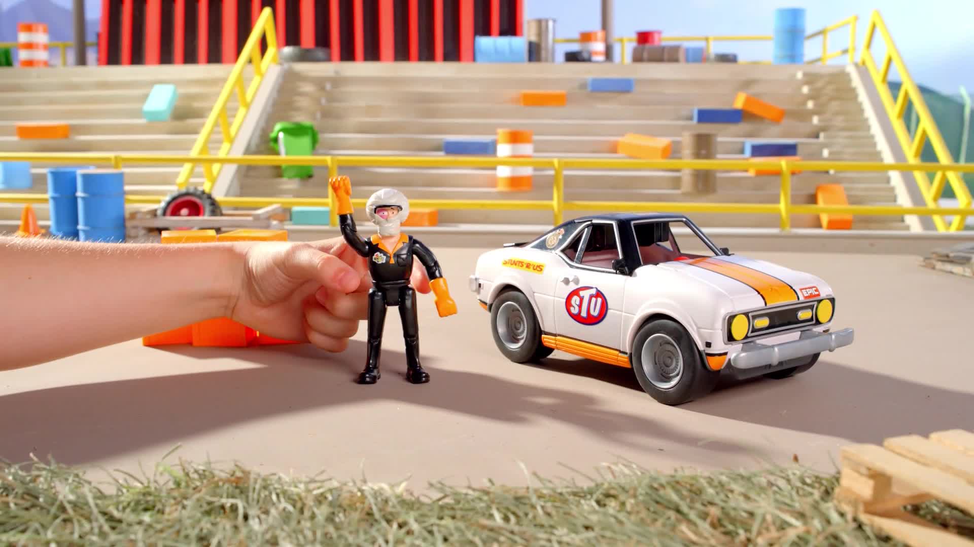 A toy race car sits next to a miniature figure, its arm raised in a signal. The figure is held by a child's hand, with a stadium backdrop behind them.
