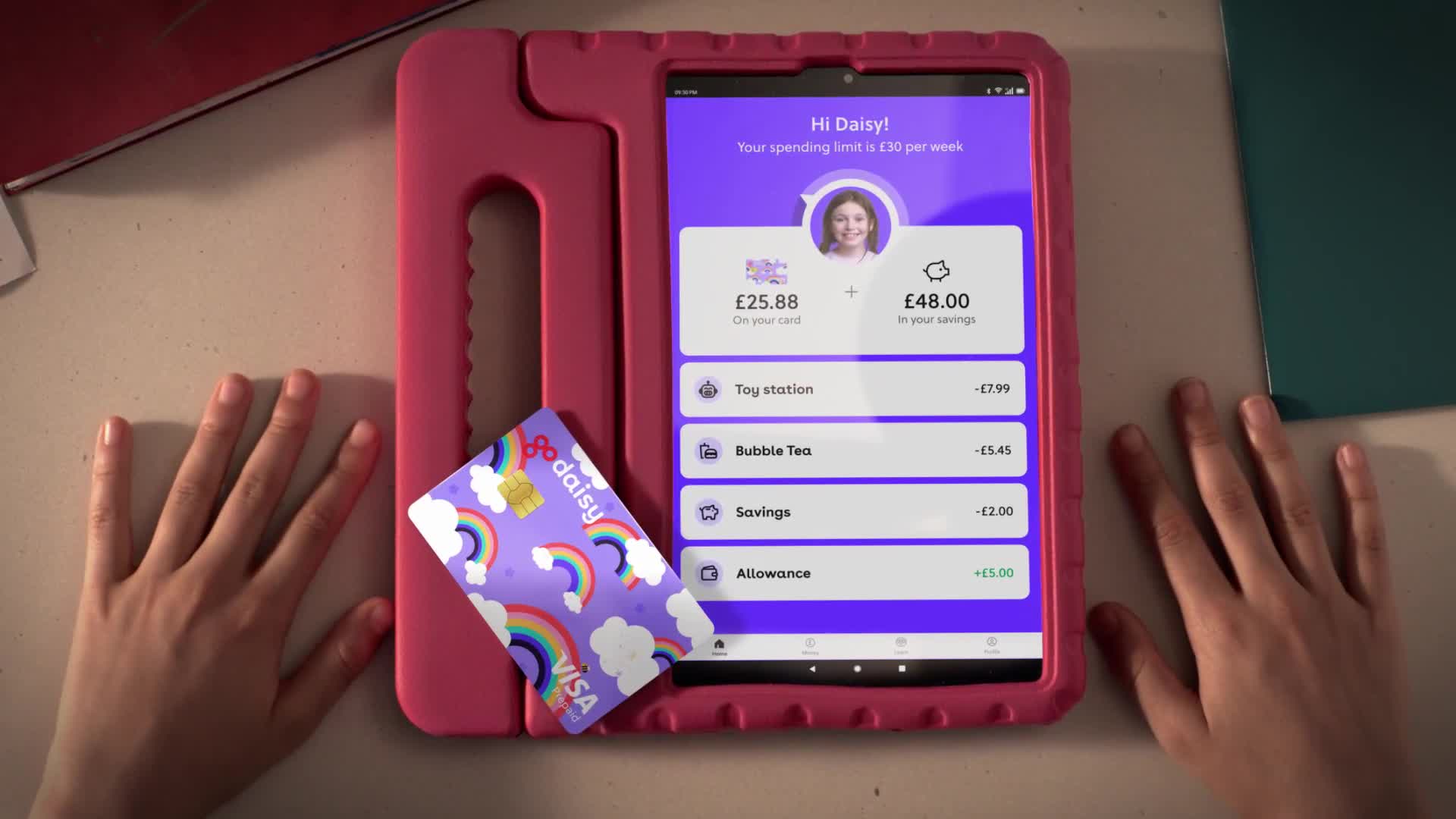 A tablet displays a UK-based financial app, showing a young girl's spending and savings. Beside it, a colorful Visa card with rainbows rests on the table.
