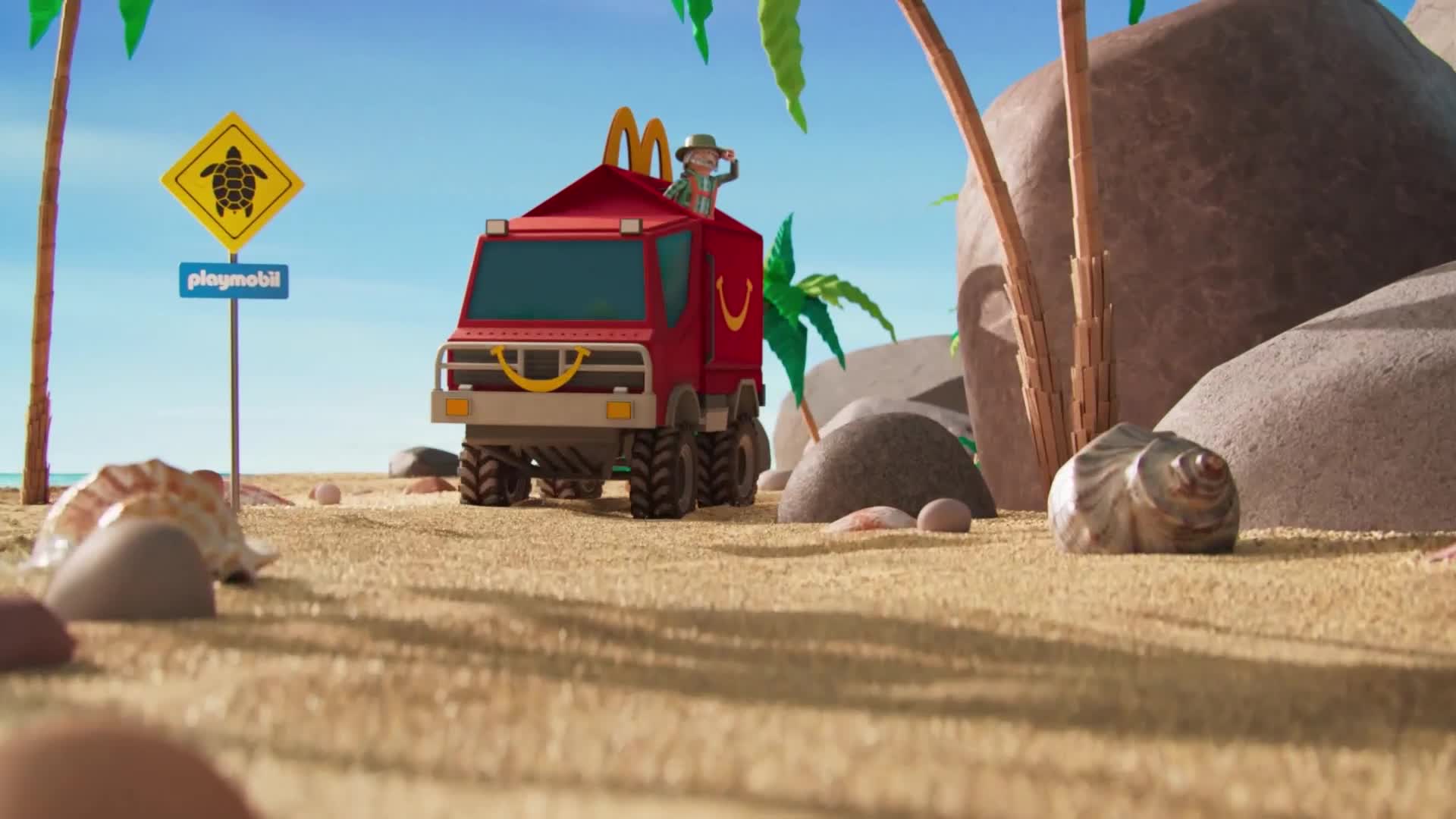 A red truck, bearing the McDonald's arches, is driving across the sandy beach. A small figure in a hat stands on top, looking ahead.
