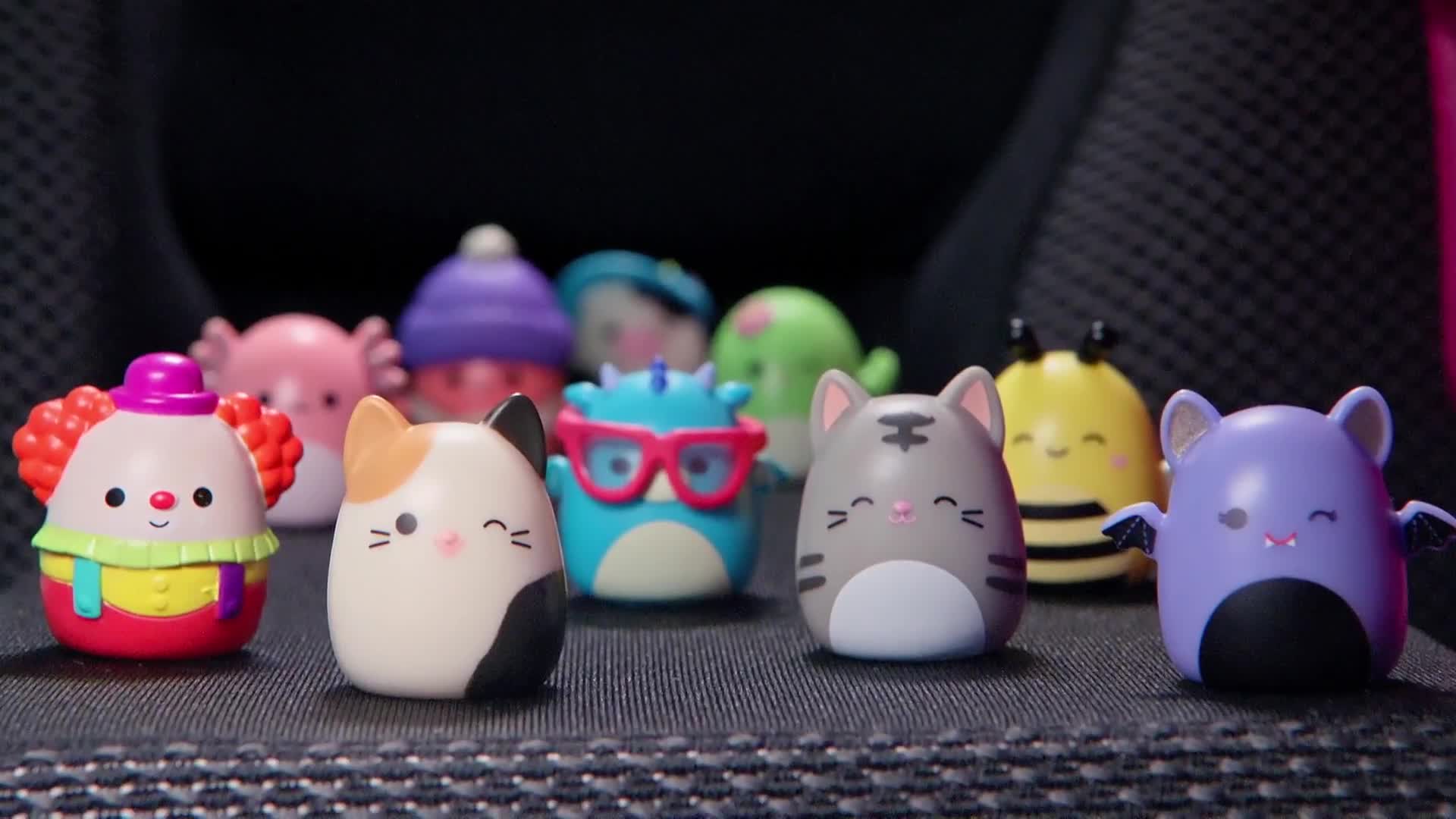 A collection of small, rounded figurines, each with a smiling face, are lined up on a textured surface. The toys, including a cat, a bee, and a clown, appear to be part of a popular pop culture trend.
