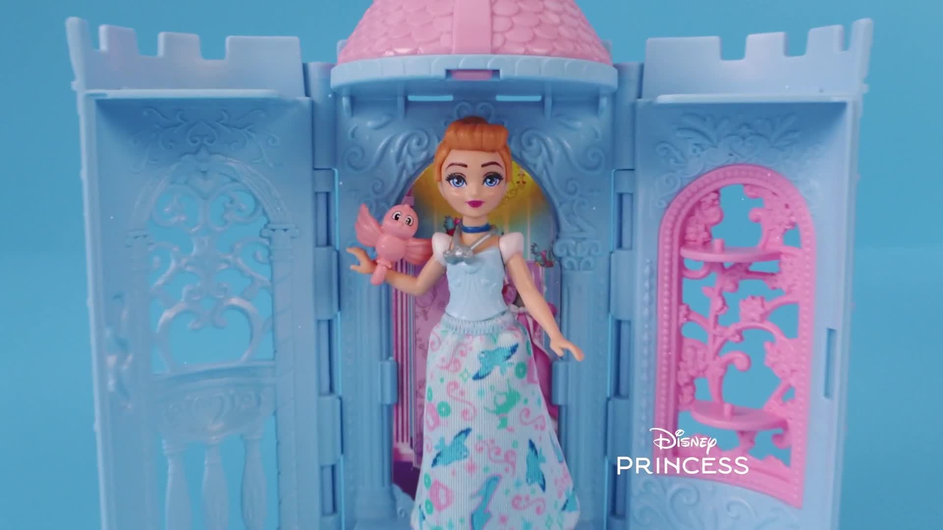 A doll, dressed in a light blue gown, stands in front of a miniature castle. She holds a small pink bird in her hand, and the Disney Princess logo is visible in the corner.
