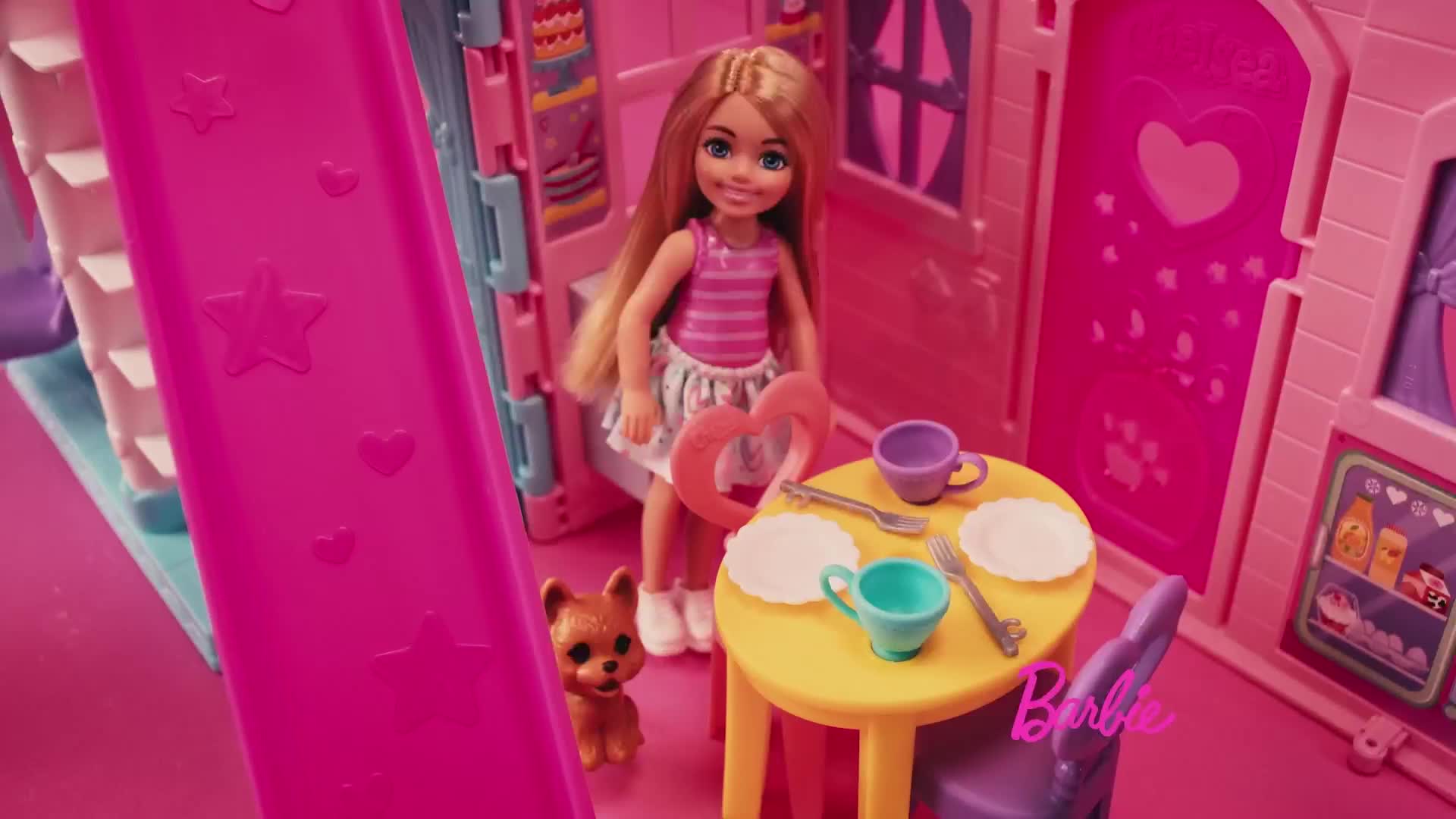 A blonde doll in a pink striped top stands in a brightly colored, miniature house. A small dog and a table set for a meal are nearby.
