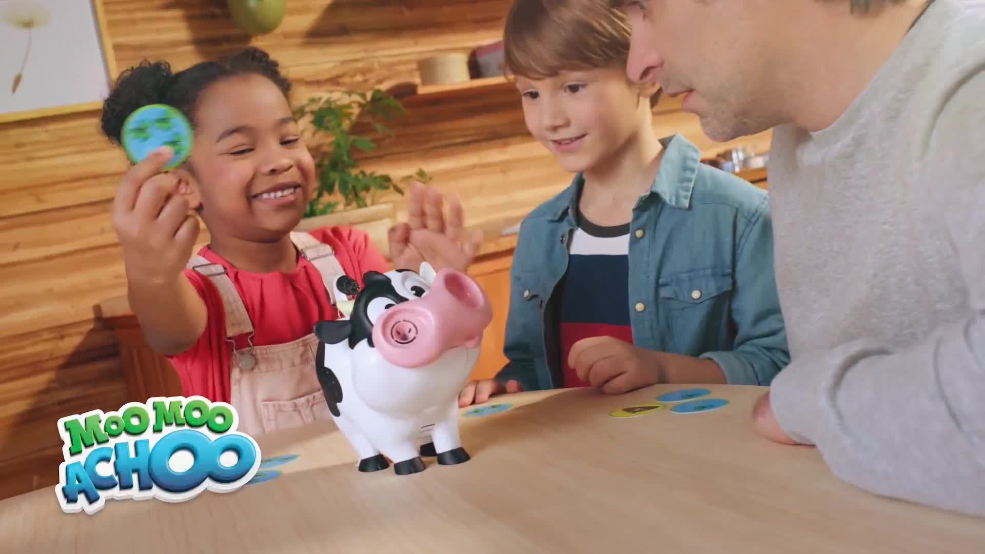 A young girl holds a small disc with a globe design, smiling at the toy cow on the table. The cow has a pink nose, and the children and an adult appear to be playing a game.
