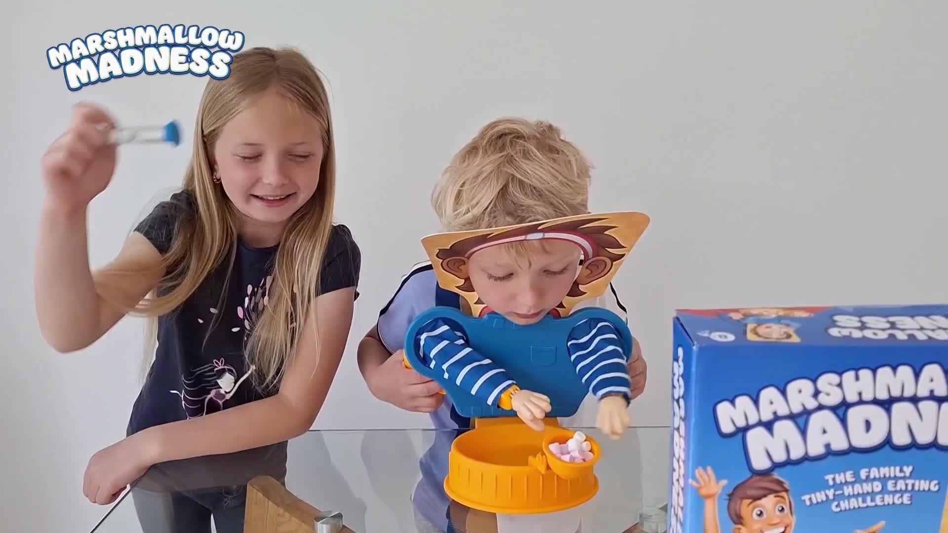 The girl is holding up a small hourglass, while the boy attempts to grab marshmallows with tiny plastic hands. A "Marshmallow Madness" game box sits on the table beside them.
