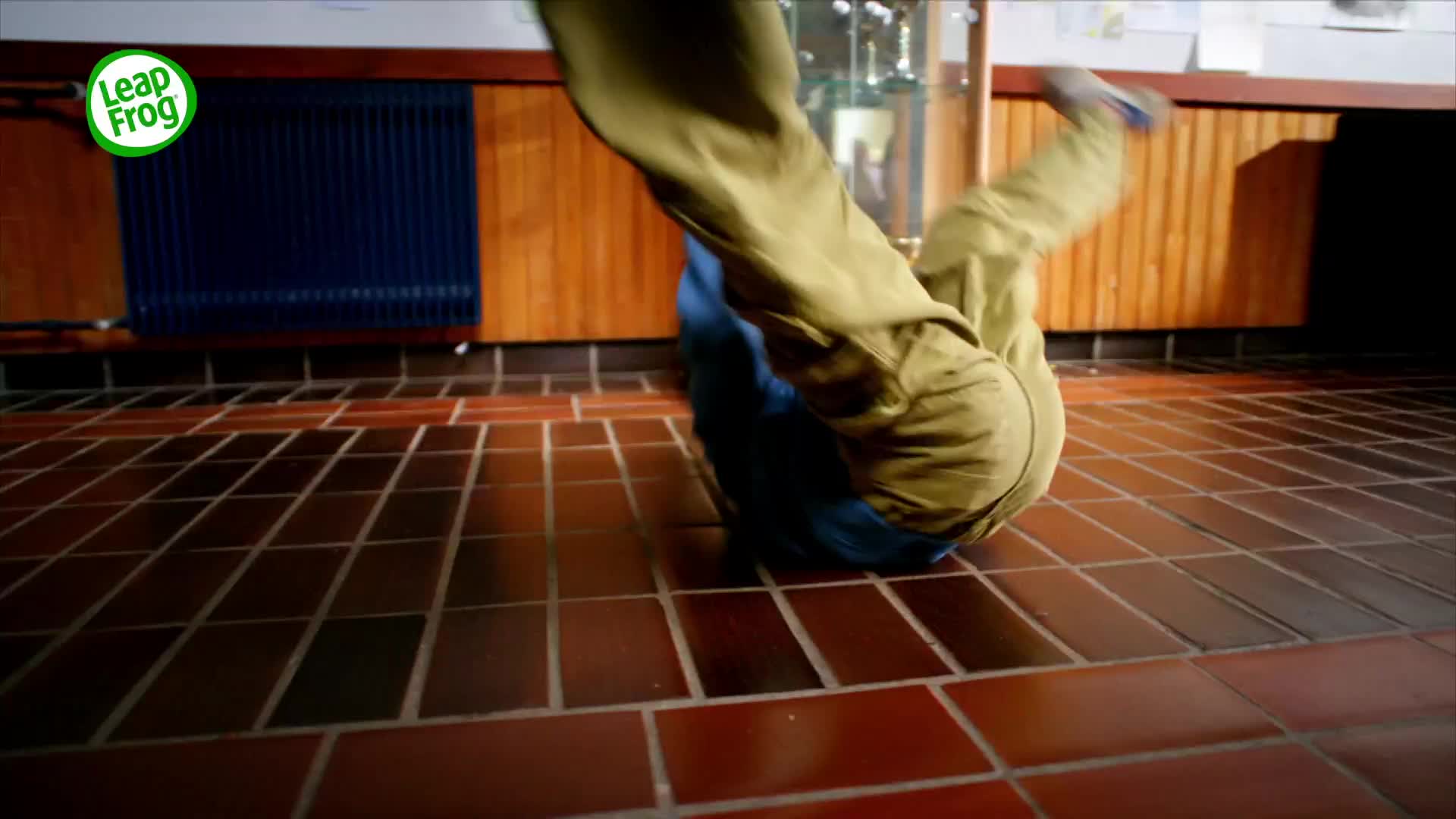 Someone is mid-somersault on the tiled floor, legs in the air. Khaki trousers and a blue shirt are visible as they tumble.
