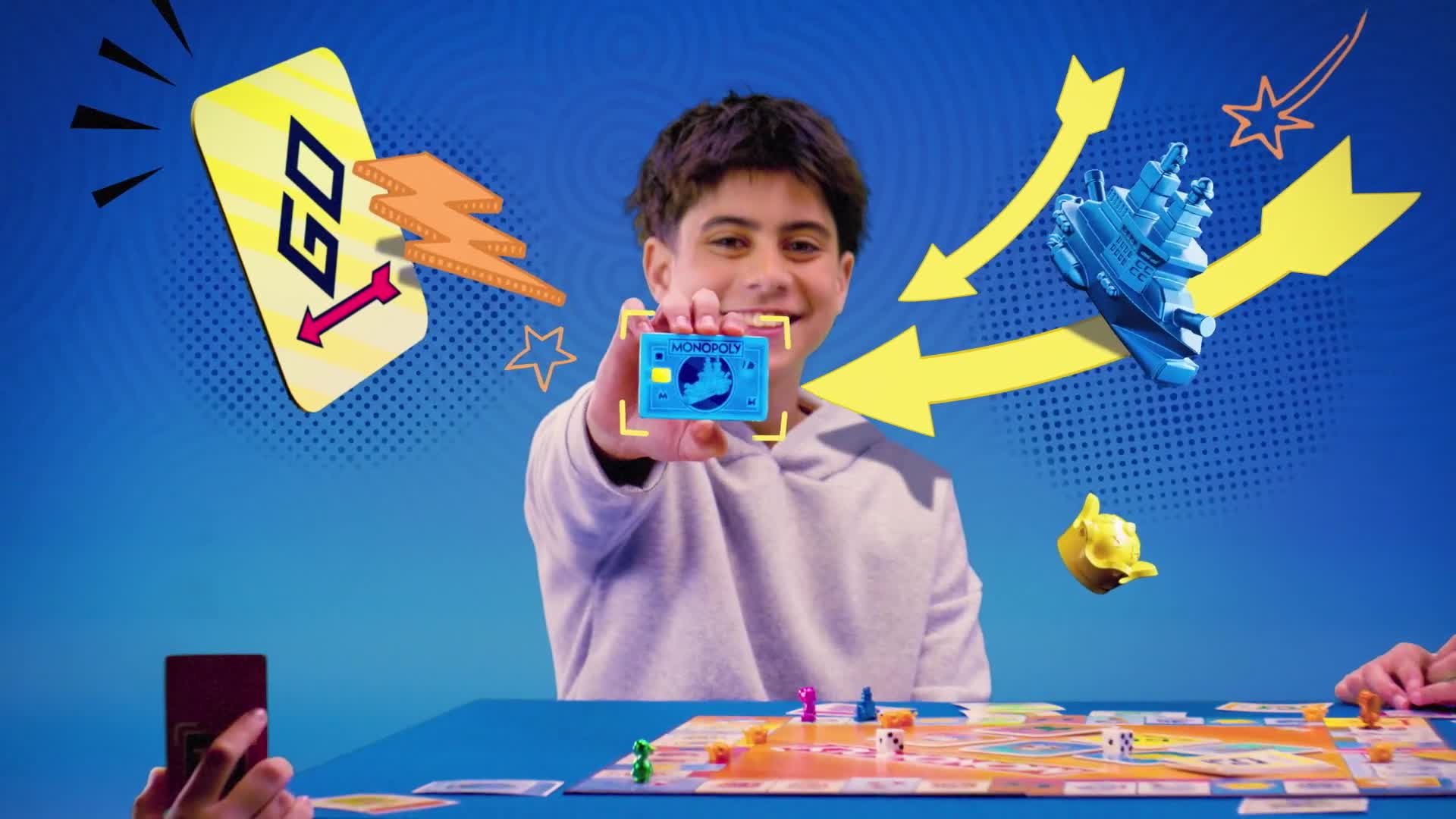 A boy grins, holding a Monopoly card towards the camera. Bright yellow arrows and comic-book style graphics surround him, while a board game sits in the foreground.
