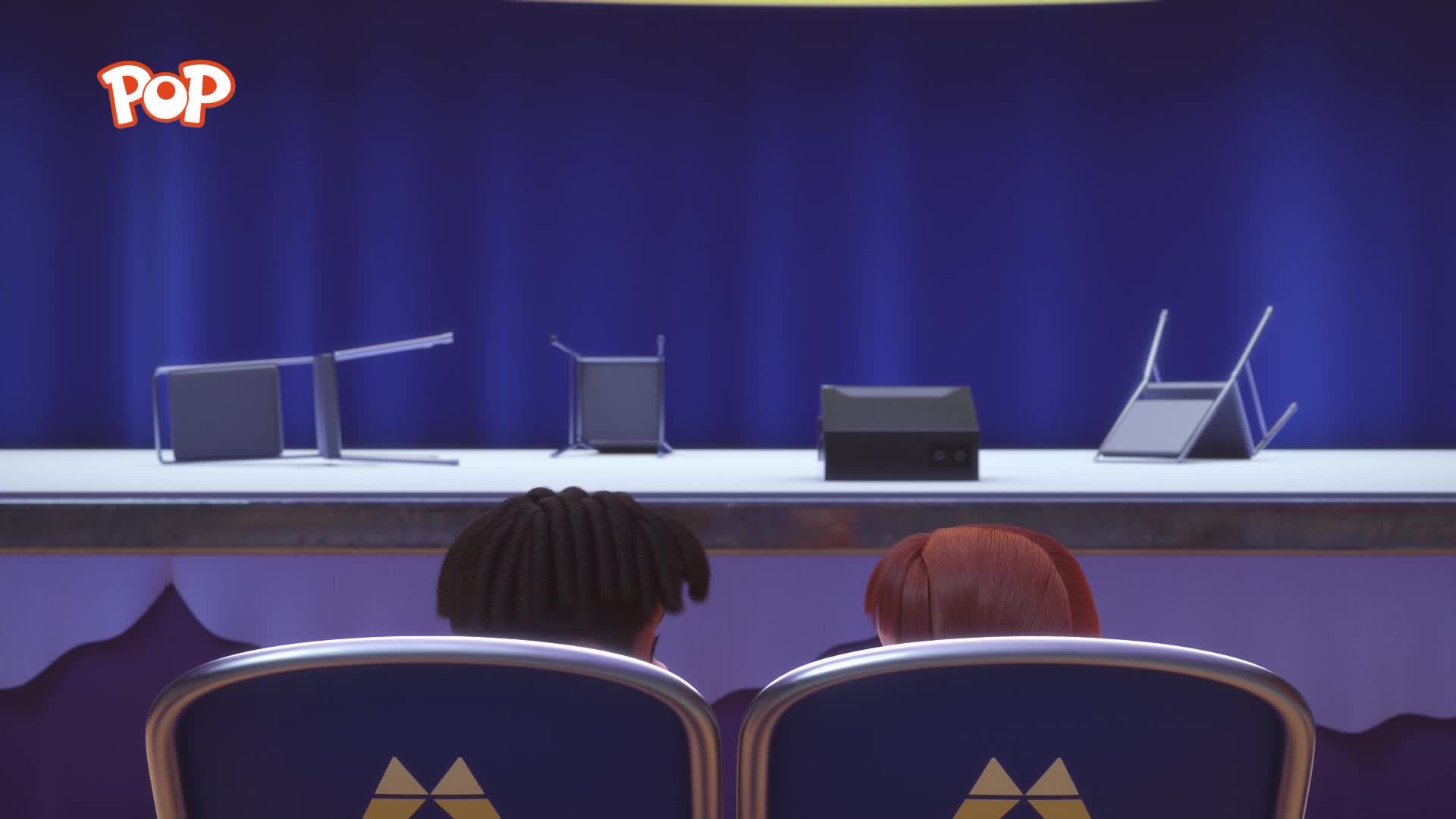 The stage is set with overturned chairs and a black box, looking like a pop performance is about to begin. Two heads, one with dark, curly hair and the other with red, are turned towards the stage from their seats.