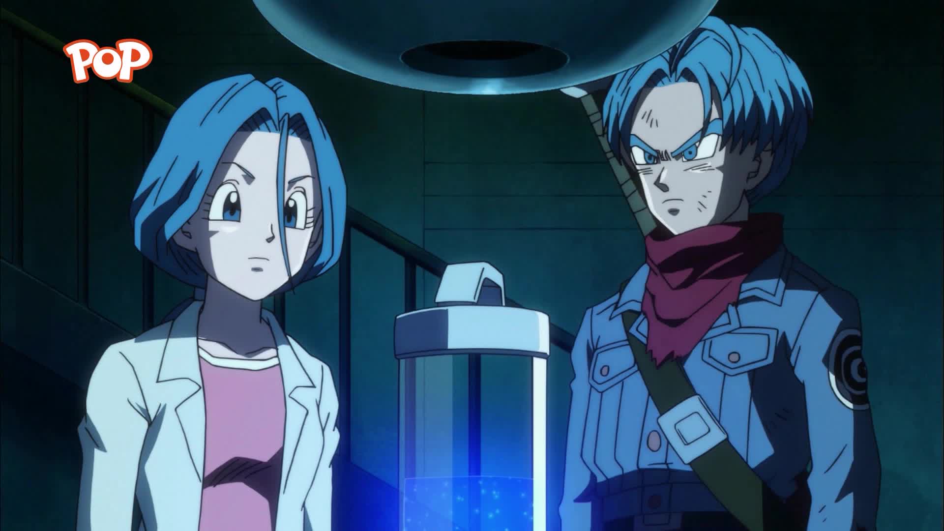 A young woman in a lab coat stands next to a young man with a red scarf. Bubbles rise in a tall glass container between them.