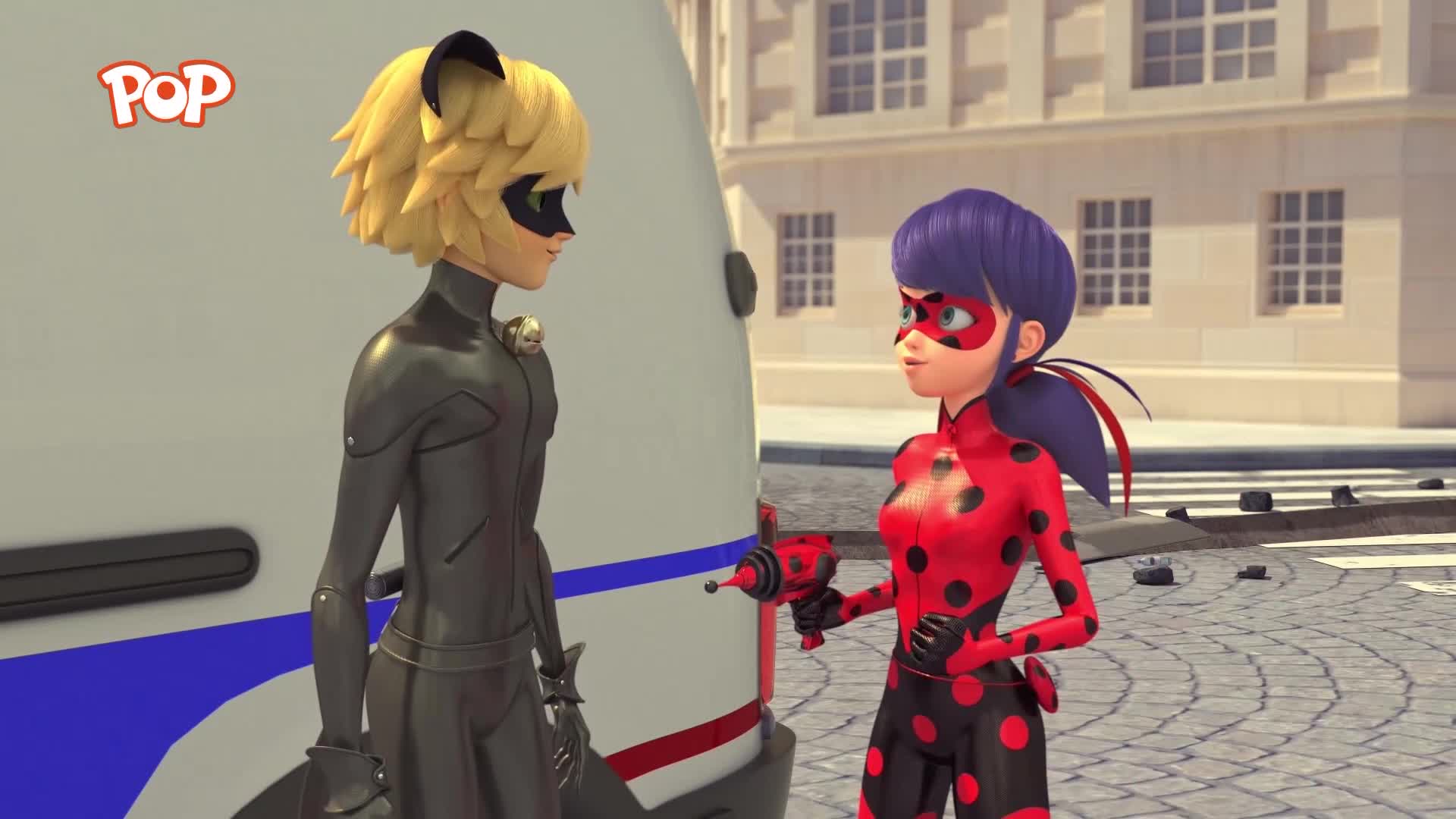 A girl in a red and black polka dot suit stands holding a device, facing a boy in a black cat-themed outfit. They are on a cobbled street, with a white van behind them.