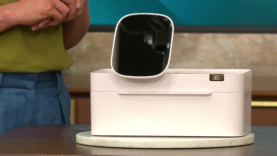 A presenter in the UK, likely on QVC Beauty, stands beside a pale pink makeup organizer, a black-mirrored panel hovering above it. The panel is angled slightly, as if about to be placed on the organizer.
