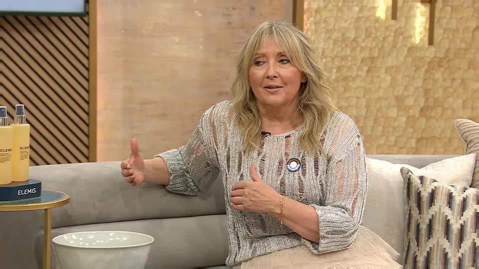 On a QVC Beauty set in the United Kingdom, a woman gestures with her hands while speaking. She's seated on a sofa, a small table to her left displaying Elemis products.
