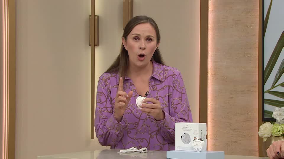The woman in the purple patterned shirt holds a small, white beauty device, pointing her index finger upwards. A product box sits on the table, matching the device she's demonstrating on the QVC Beauty show from the UK.
