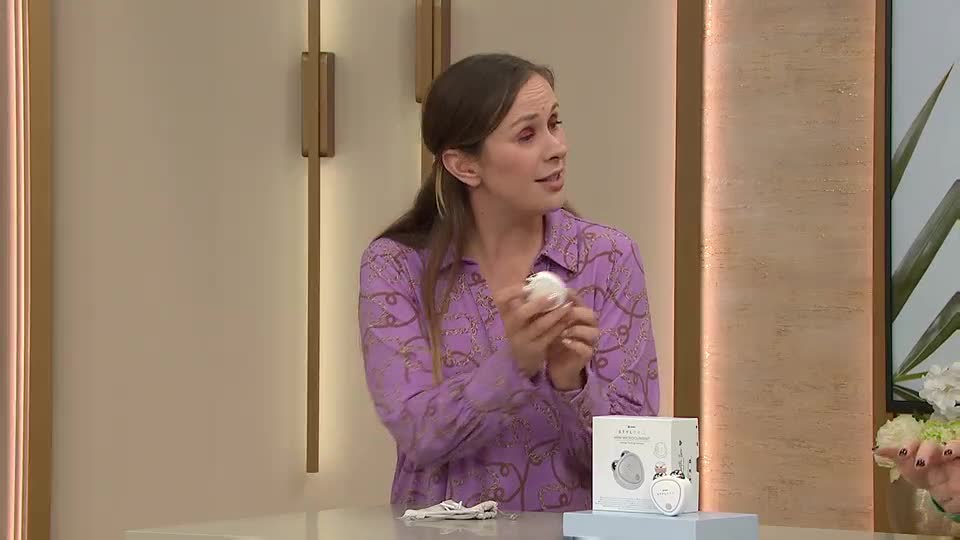 A woman in a patterned purple shirt holds a small, white beauty product, gesturing as she speaks. Beside her, a box and another product of the same kind sit on a display, suggesting a QVC Beauty presentation.
