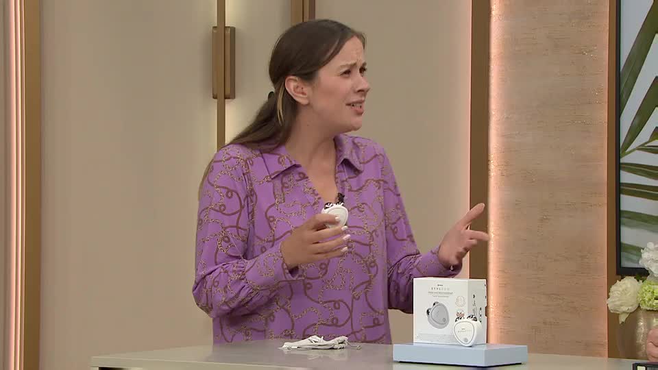 A woman in a patterned purple shirt gestures emphatically while holding a small, white device. A box with the same device sits on a table, along with a small, discarded cloth.
