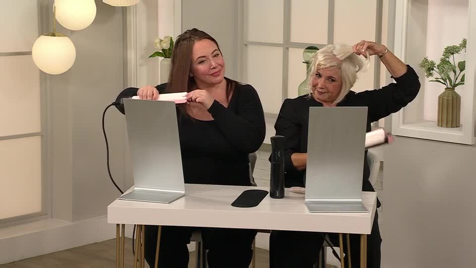 Two women, each with a flat iron, are demonstrating hair styling techniques. One woman is smoothing her hair, while the other lifts her hand to show off the volume of her hair.
