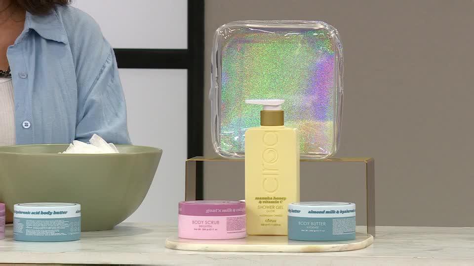 On a QVC Beauty set in the United Kingdom, a woman stands near a large bowl of what looks like whipped cream. Various body care products are arranged on a small platform nearby, including a yellow shower gel and a pink body scrub.
