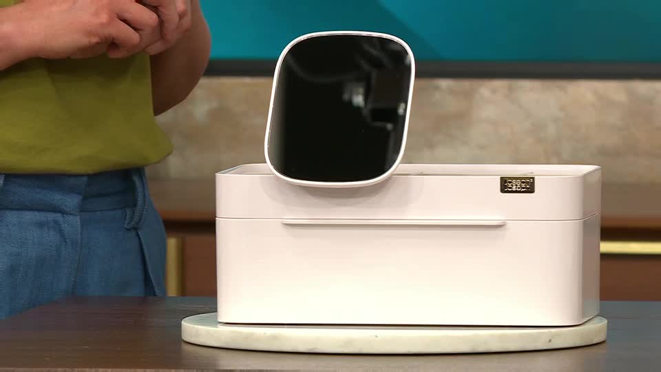A presenter in QVC Beauty, United Kingdom, is demonstrating a white beauty organizer with a sleek, black mirror attached. The mirror is angled slightly above the box, and the presenter's hands are visible in the background.
