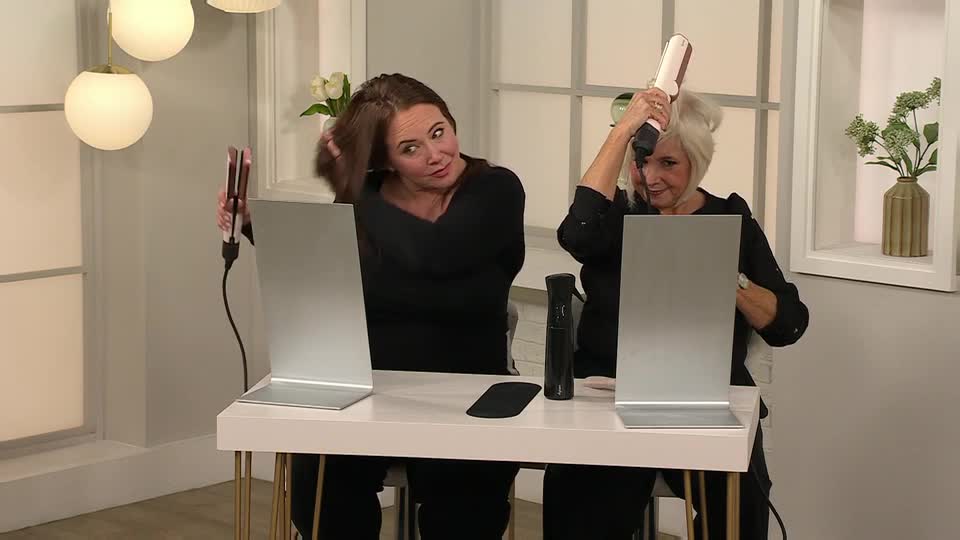 Two women in black tops are demonstrating hair styling products, each holding a flat iron. They're positioned behind a white table, with a mirror and a spray bottle in front of each of them.
