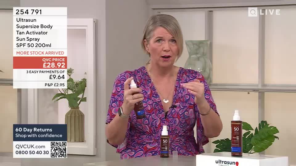 A woman on QVC in the United Kingdom holds a bottle of sun spray, gesturing with her other hand as she talks. Two more bottles sit on the table in front of her, and the price is displayed on a screen.
