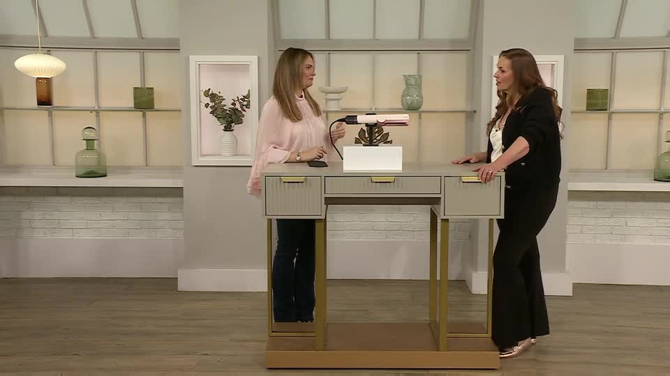 Two women stand behind a modern desk, one holding a small device, the other gesturing as they talk. A hair styling tool sits on a white box atop the desk, ready for demonstration.
