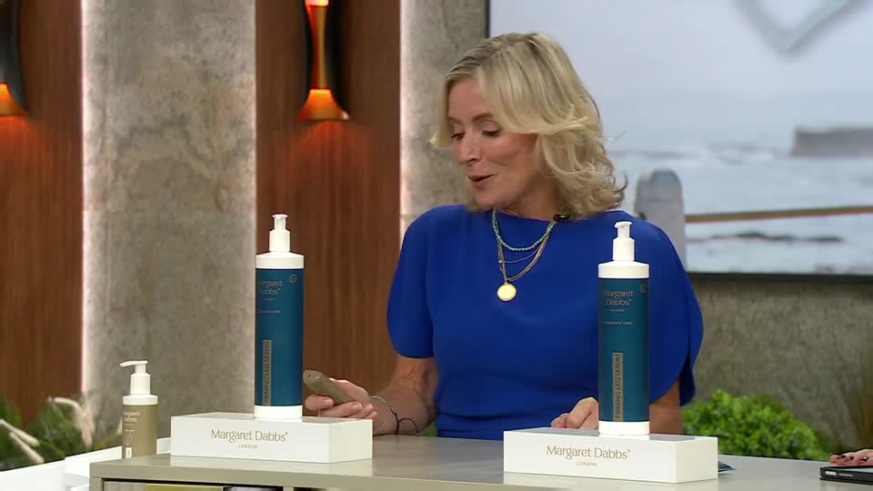 A woman in a blue top is holding a small, brown item while presenting Margaret Dabbs London products on QVC Beauty. Two tall, blue bottles with white pumps sit on white boxes in front of her.

