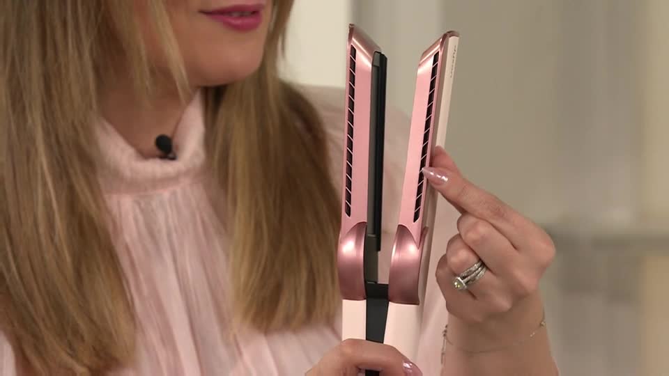A woman, wearing a pink top, is holding a rose gold hair straightener. She's pointing to the side of the device, likely demonstrating a feature on QVC Beauty.
