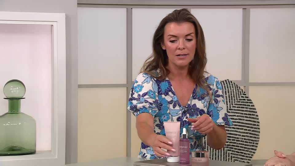 The woman in the floral dress gestures towards a selection of beauty products, holding them up for the camera. She's speaking, presumably about the creams and serums lined up on the table in front of her.
