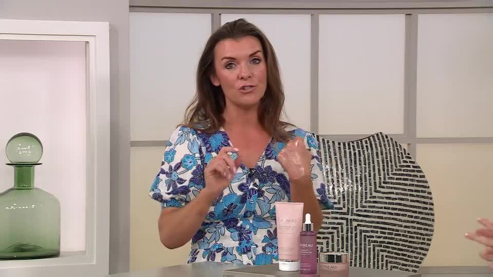 A woman in a floral dress gestures with her hands, demonstrating a skincare product for the QVC Beauty audience. She holds her hand up, showing the product on her skin while speaking.
