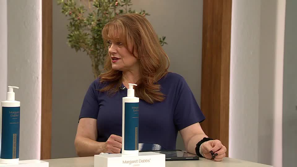 A woman in a navy top gestures towards a bottle of lotion on a white stand. The QVC Beauty presenter is speaking, presumably about the product.
